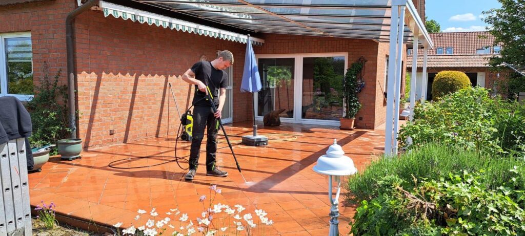Marius Steinert reinigt eine mit Terrakotta-Fliesen belegte Terrasse mit einem Hochdruckreiniger. Die Terrasse gehört zu einem roten Backsteinhaus mit Glasüberdachung und ist von grünen Pflanzen umgeben.